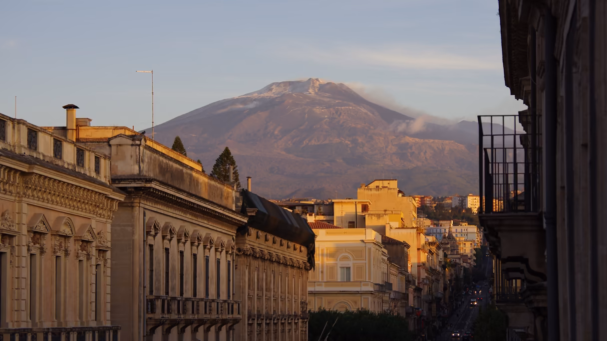 El Etna al atardecer visto desde una calle de Catania: la montaña ancha y oscura con nieve en la cumbre y una nube de humo, y delante, tejados y fachadas de palacios iluminados por el sol bajo.