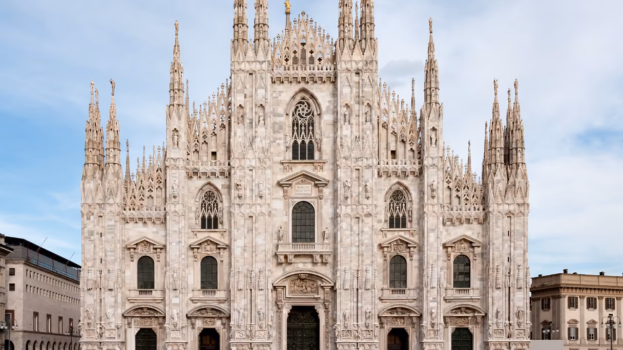 La fachada del Duomo de Milán de frente, de mármol claro, bajo un cielo azul con nubes: agujas y pináculos con estatuas, un ventanal gótico en el centro y ventanas y puertas de bronce entre columnas labradas; a los lados, los edificios de la plaza.