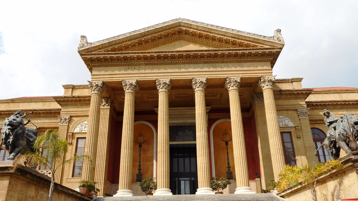 El Teatro Massimo de Palermo de frente: un pórtico de seis columnas corintias de piedra dorada bajo un frontón triangular, puertas altas entre muros rojos y, a los lados, dos leones de bronce con figuras sentadas; abajo, el arranque de la escalinata.