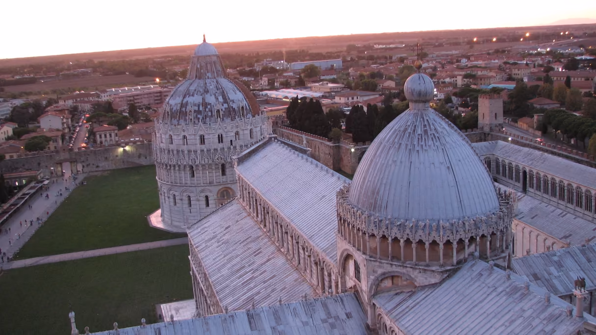El baptisterio y la catedral de Pisa vistos desde lo alto de la torre al atardecer: la cúpula gris del baptisterio a la izquierda, sobre el césped de la plaza, y en primer plano los tejados de plomo de la catedral con su cúpula; al fondo, la ciudad y el campo bajo un cielo rosado.