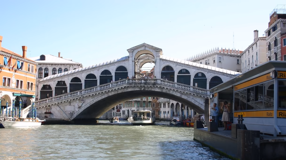 El puente de Rialto de Venecia visto desde el Gran Canal: el arco de piedra blanca con su pórtico central y las dos filas de arcadas, un vaporetto pasando por debajo y, a la derecha, la parada de vaporetto que lleva el nombre de Rialto.