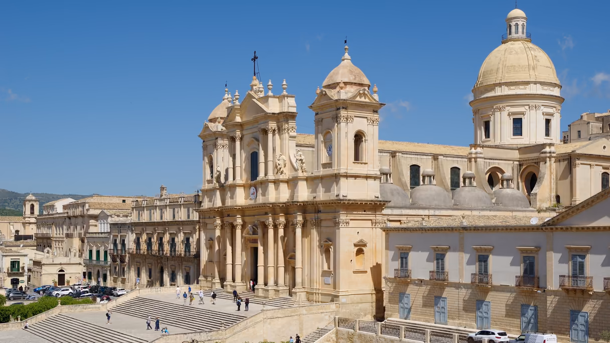 La catedral de Noto desde lo alto: una fachada barroca de piedra dorada con dos campanarios, sobre una escalinata ancha con gente pequeña; a la derecha, la cúpula, y alrededor, palacios del mismo color bajo un cielo azul.