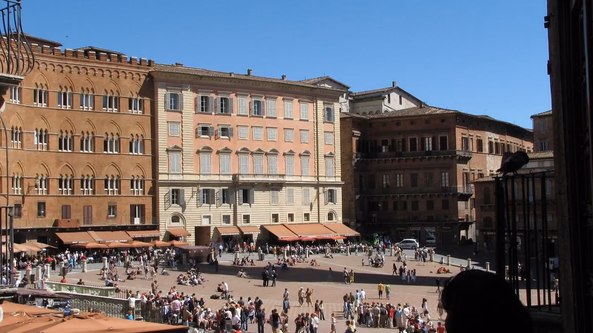 La Piazza del Campo de Siena a mediodía, desde lo alto de uno de sus bordes: palacios de ladrillo y de color salmón con toldos y terrazas, y la plaza de ladrillo en pendiente llena de gente paseando y sentada en el suelo.