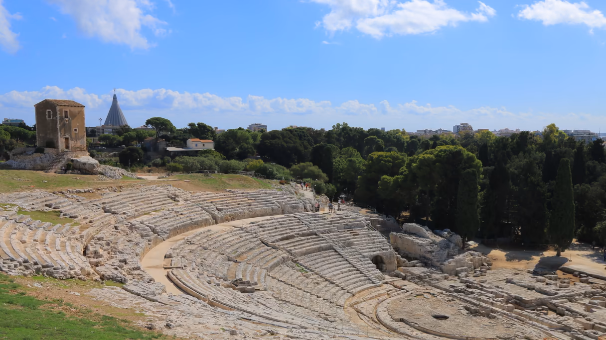 El teatro griego de Siracusa visto desde lo alto: las gradas de piedra clara excavadas en la roca en un gran semicírculo, una casa de piedra en lo alto de las gradas, un bosque de pinos y cipreses detrás y un cielo azul con nubes.