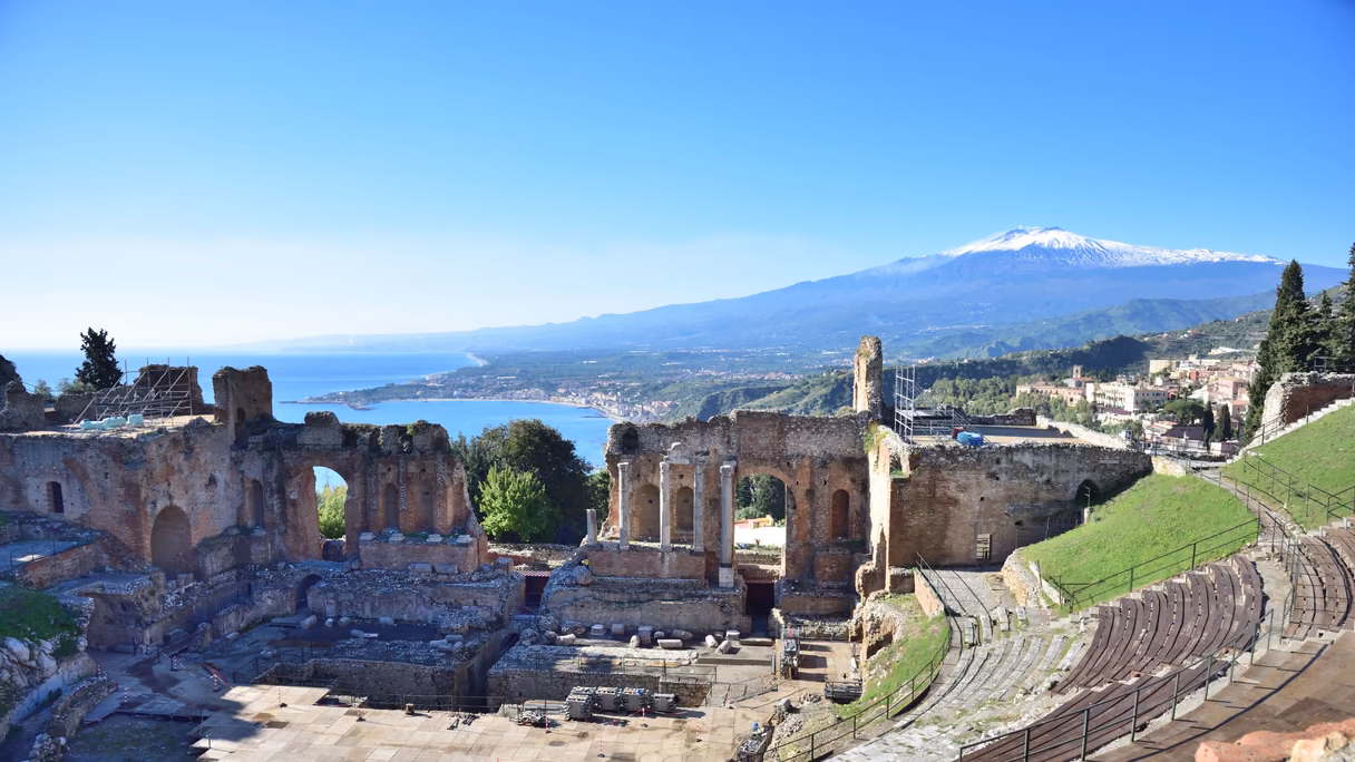 El teatro antiguo de Taormina desde lo alto de sus gradas: ruinas de ladrillo con arcos y columnas, la costa y el mar azul a la izquierda y, al fondo, el Etna con la cumbre nevada bajo un cielo despejado.