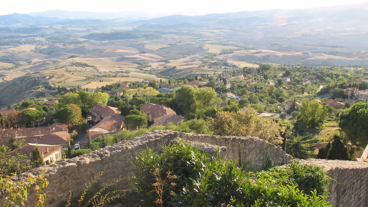 Las colinas de la Toscana desde la muralla de Volterra a media tarde: en primer plano, el muro de piedra con matorral; debajo, casas de tejado rojo entre árboles, y al fondo, campos ondulados de tonos ocres y verdes hasta los montes.