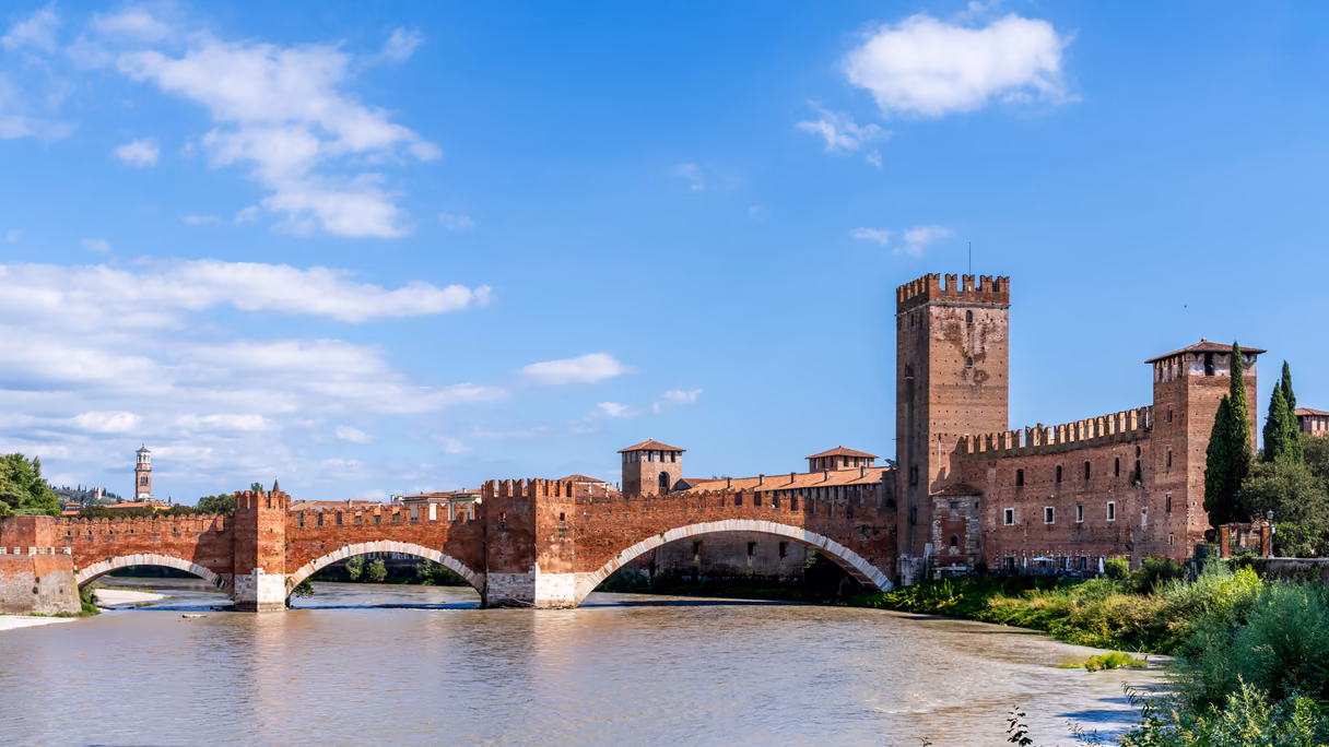 Castelvecchio de Verona y su puente sobre el Adigio bajo un cielo azul con nubes: el puente de ladrillo rojo con almenas y tres arcos sobre el río, el castillo con su torre alta a la derecha y, a lo lejos a la izquierda, la torre de los Lamberti.